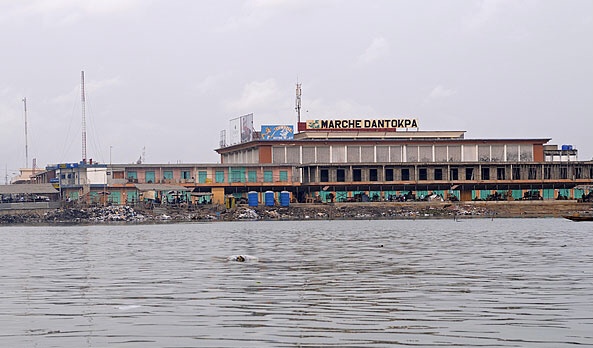 grand bâtiment long vu de loin avec panneau marché dantokpa face à un plan d'eau calme sous un ciel nuageux