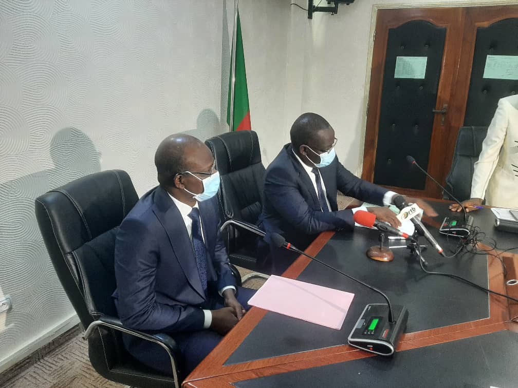 Deux hommes en costume et masque assis à une table avec microphones et documents dans une salle de réunion