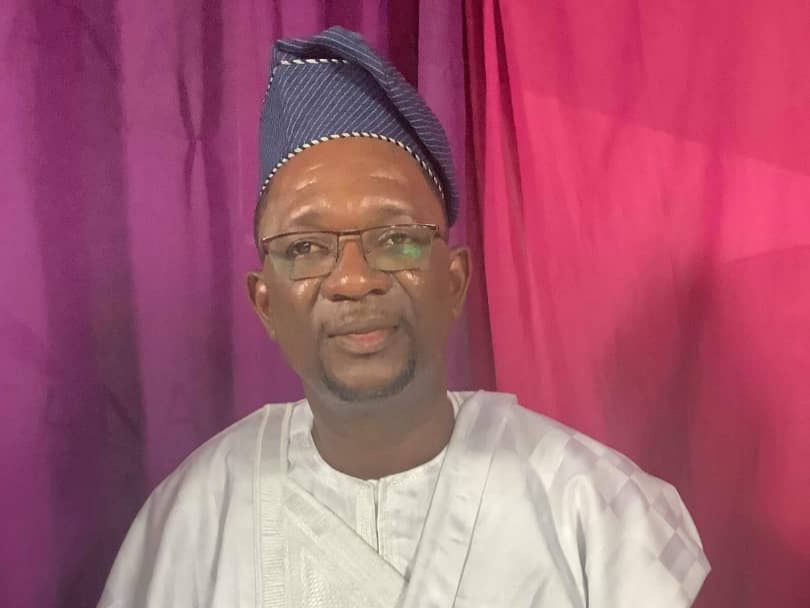 Homme portant un boubou blanc et un chapeau traditionnel assis devant un rideau rouge et violet