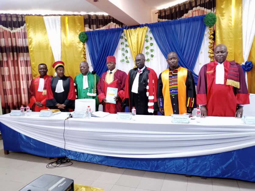 Sept hommes en toges officielles debout derrière une table drapée blanc, bleu et or dans une salle ornée de rideaux colorés