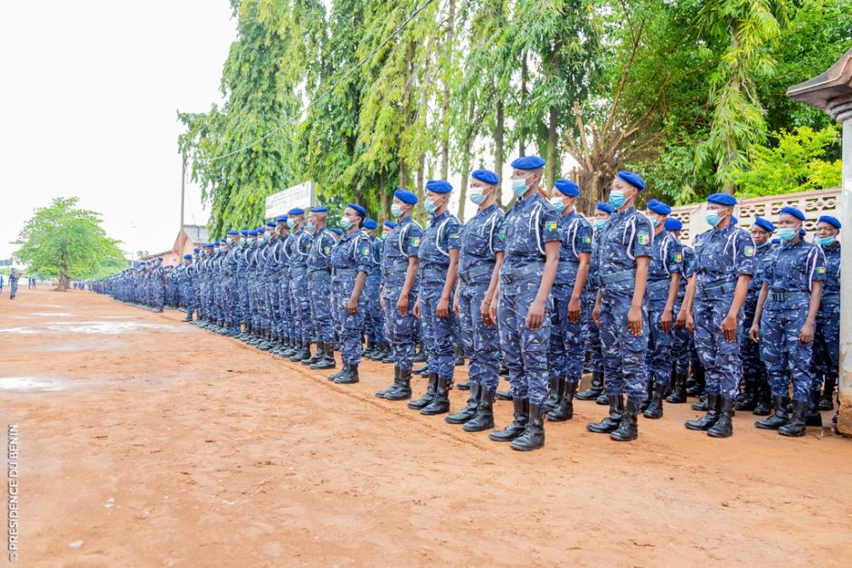 Une longue file d’agents en uniforme camouflage bleu avec casquettes bleues alignés sur un sol en terre battue et arbres en arrière-plan