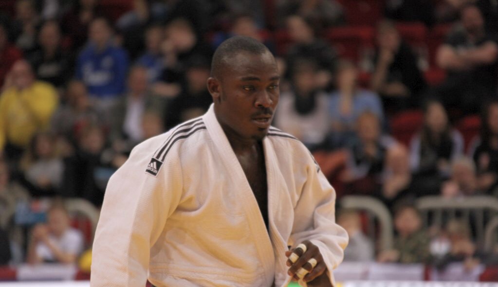Judoka masculin en kimono blanc en action sur un tatami devant un public flou en arrière-plan