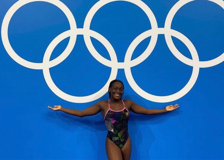 Une nageuse souriante en maillot de bain debout devant un mur bleu avec les anneaux olympiques blancs