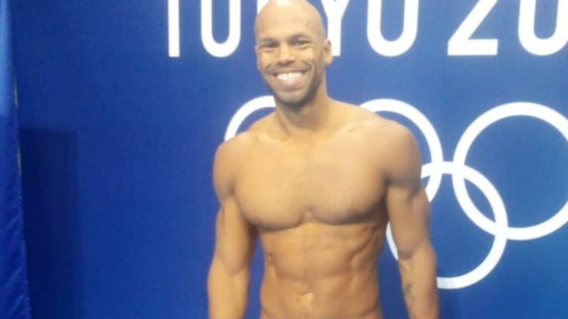 Homme souriant en maillot de bain devant un mur bleu avec logo des Jeux Olympiques de Tokyo 2020