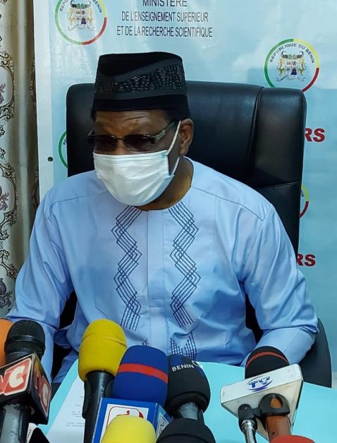 Homme portant une tenue traditionnelle bleue, un masque blanc et des lunettes assis face à plusieurs microphones devant un panneau officiel.