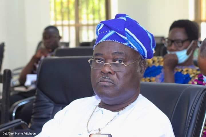 Homme âgé portant un bonnet traditionnel bleu et des lunettes, assis lors d'une réunion avec plusieurs personnes en arrière-plan