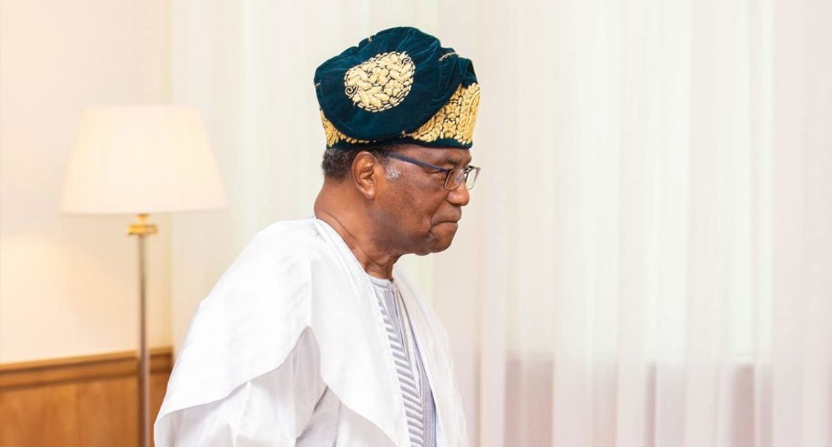 Homme âgé portant une tenue traditionnelle blanche et un bonnet orné de motifs dorés debout près d'un rideau blanc translucide