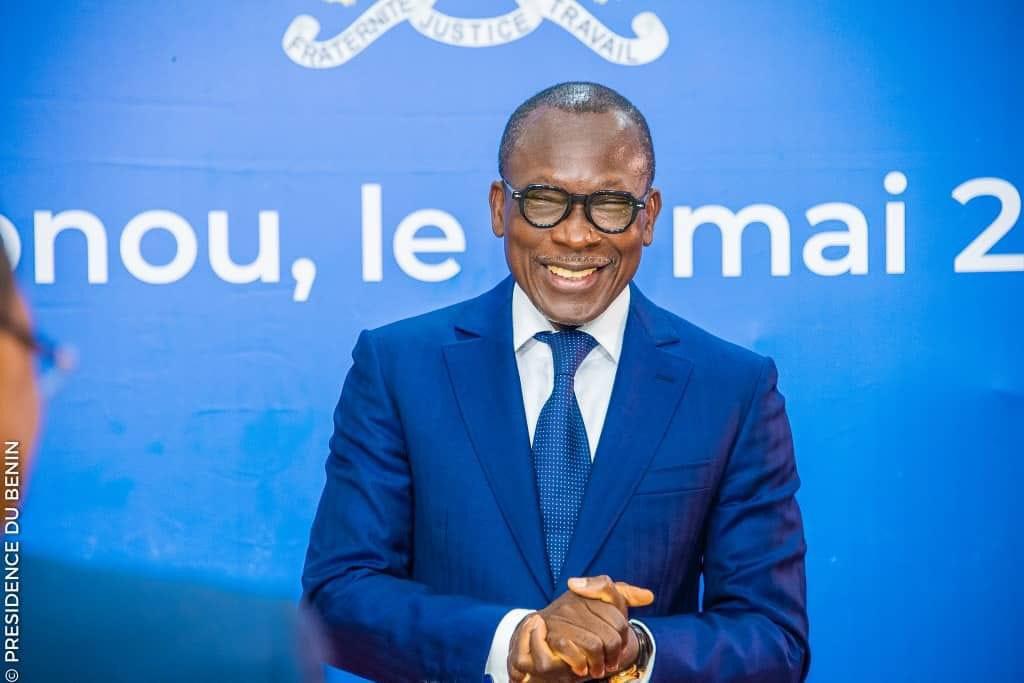 Un homme en costume bleu sourit devant un fond bleu avec du texte partiellement visible