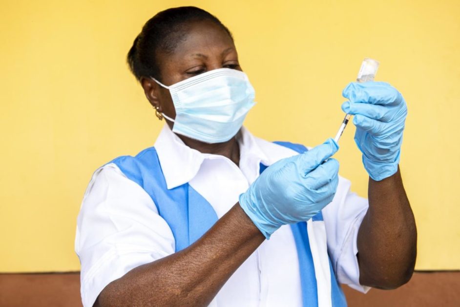 Une professionnelle de santé masquée et gantée prépare une seringue avec un vaccin devant un mur jaune