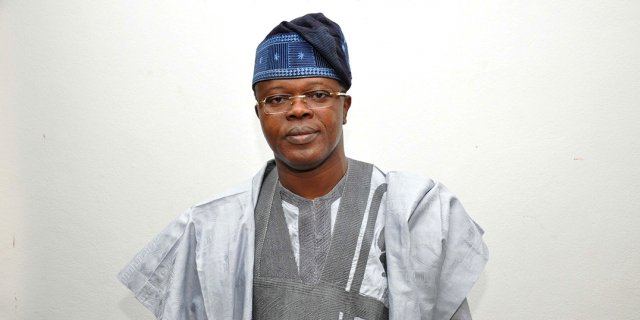 Homme portant un boubou gris clair et un bonnet traditionnel bleu pose devant un mur blanc