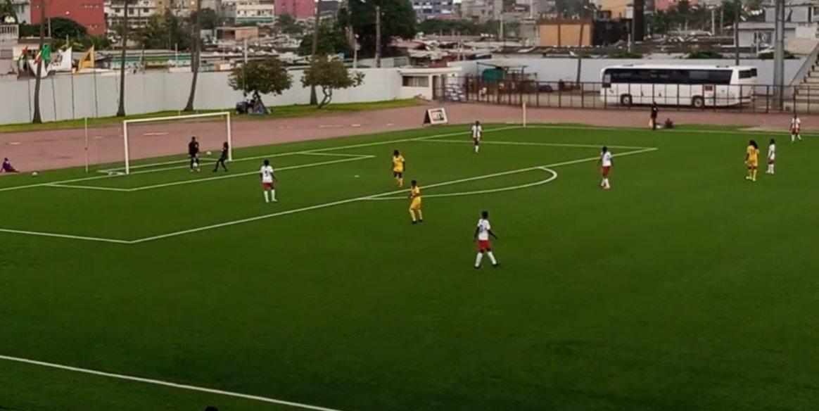 Dix joueuses en action sur un terrain de football vert entouré de bâtiments urbains et d’un bus blanc stationné à l’arrière-plan