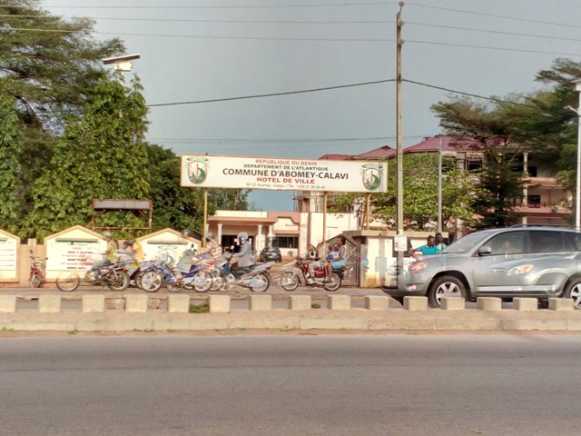 Entrée d’un bâtiment administratif avec plusieurs motos garées et des personnes présentes au niveau du portail visible depuis la route principale
