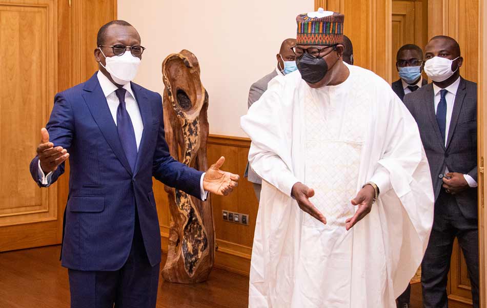 Deux hommes en discussion portent des masques, un en costume bleu et l'autre en tenue blanche traditionnelle, d'autres personnes en costume sont en arrière-plan