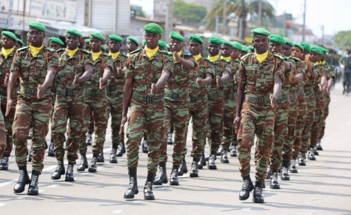 Des militaires en uniforme camouflé et béret vert défilent en formation serrée sur une rue urbaine