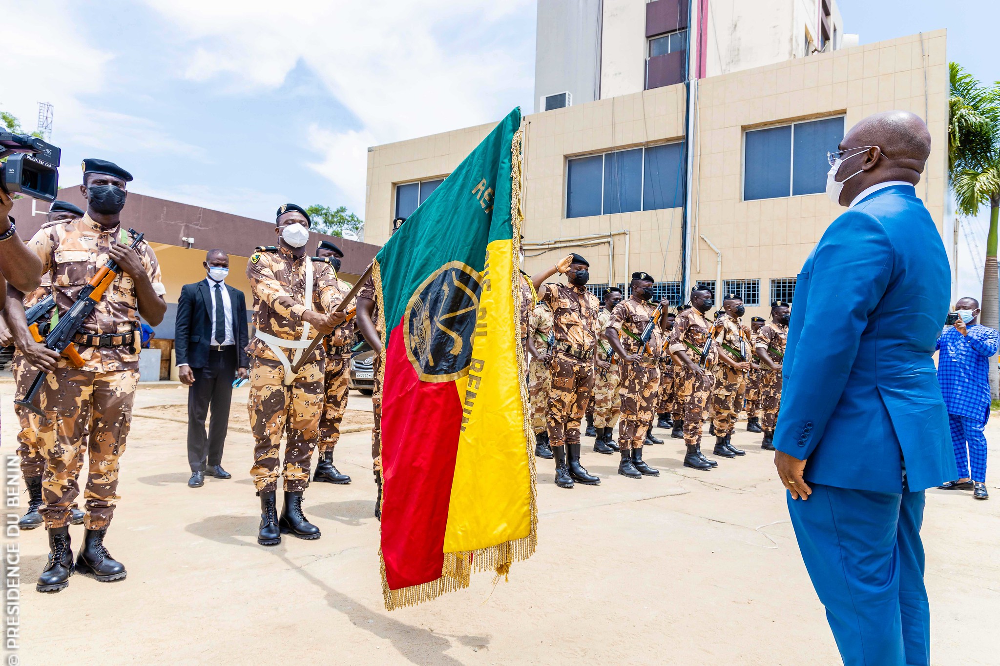 Des soldats en uniforme camouflage, certains armés, en formation devant un homme en costume bleu et masque blanc devant un bâtiment moderne