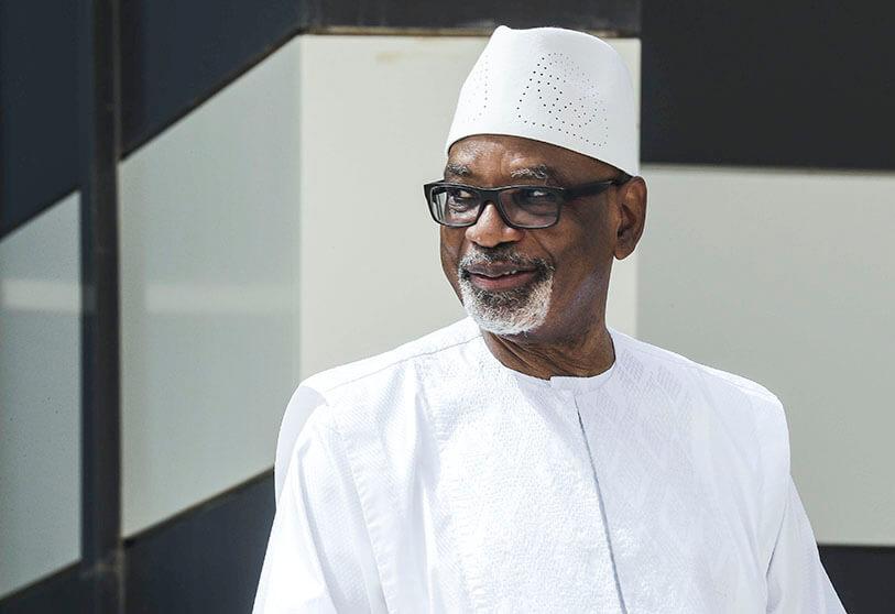 Homme souriant portant une tenue traditionnelle blanche et un chapeau assorti devant un fond aux formes géométriques noires et blanches