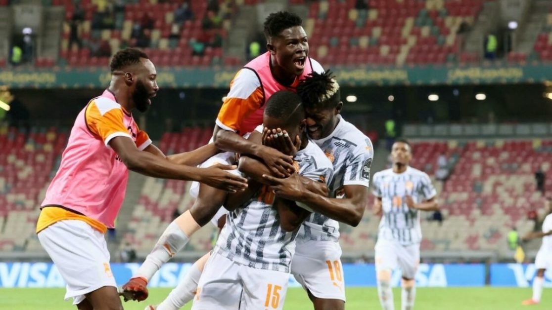 Des joueurs de football en maillots blancs et orange célèbrent un but collectif dans un stade avec tribunes partiellement vides