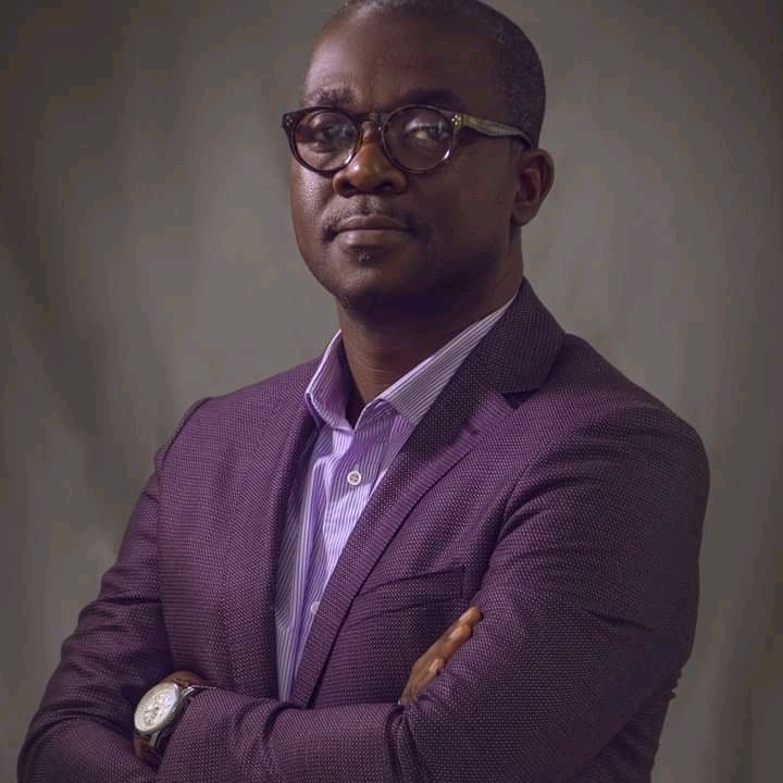 Homme portant des lunettes, veste violette et chemise, bras croisés, regardant l’objectif, fond neutre flou