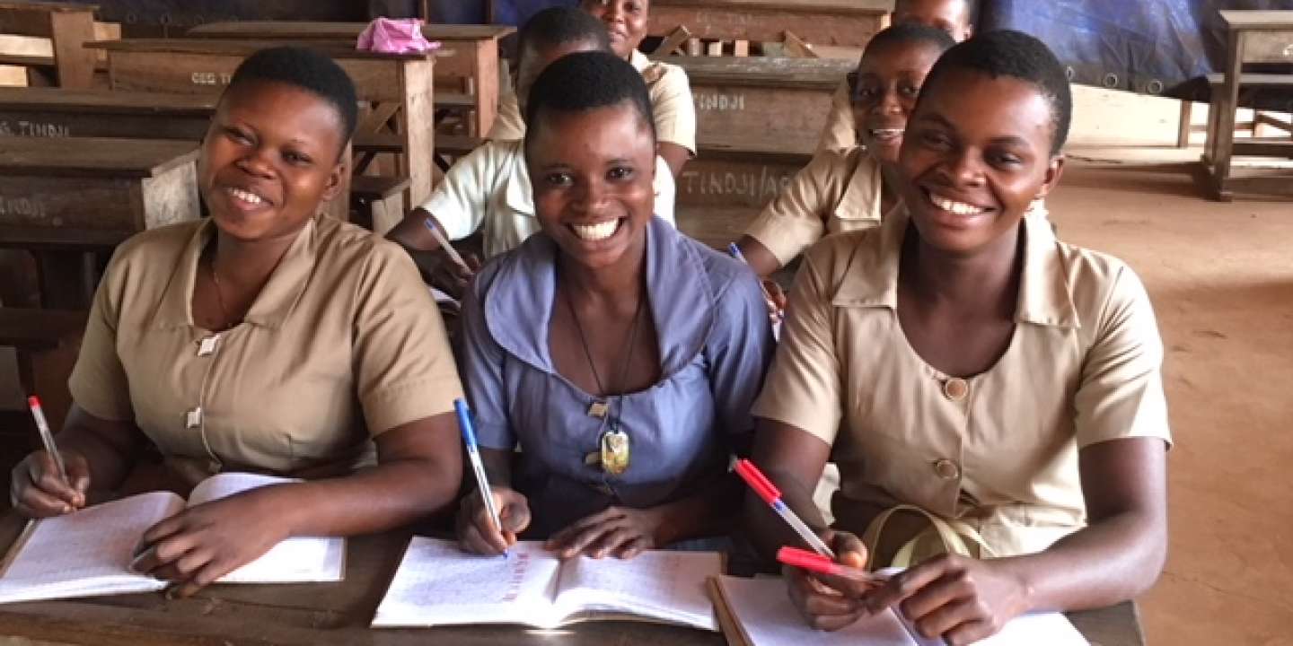 Trois jeunes filles souriantes en uniforme écrivent dans leurs cahiers assises dans une salle de classe en bois