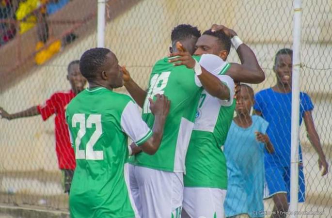 Trois joueurs en maillots verts et blancs s’embrassent et célèbrent un but devant un but de football avec des enfants en arrière-plan