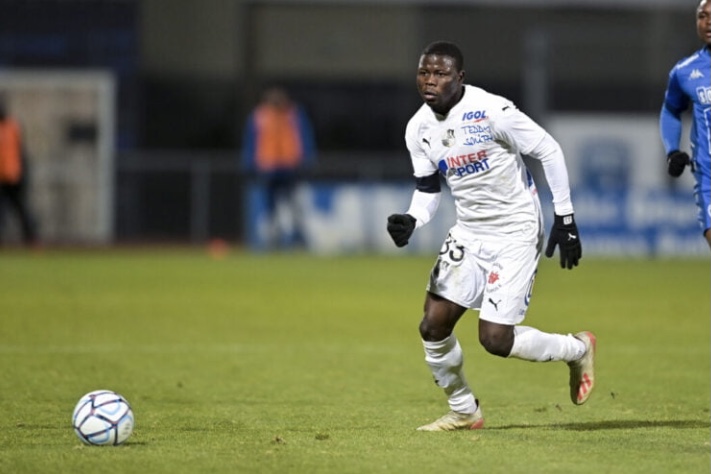 Un joueur en tenue blanche en mouvement sur un terrain de football avec un ballon au sol devant lui
