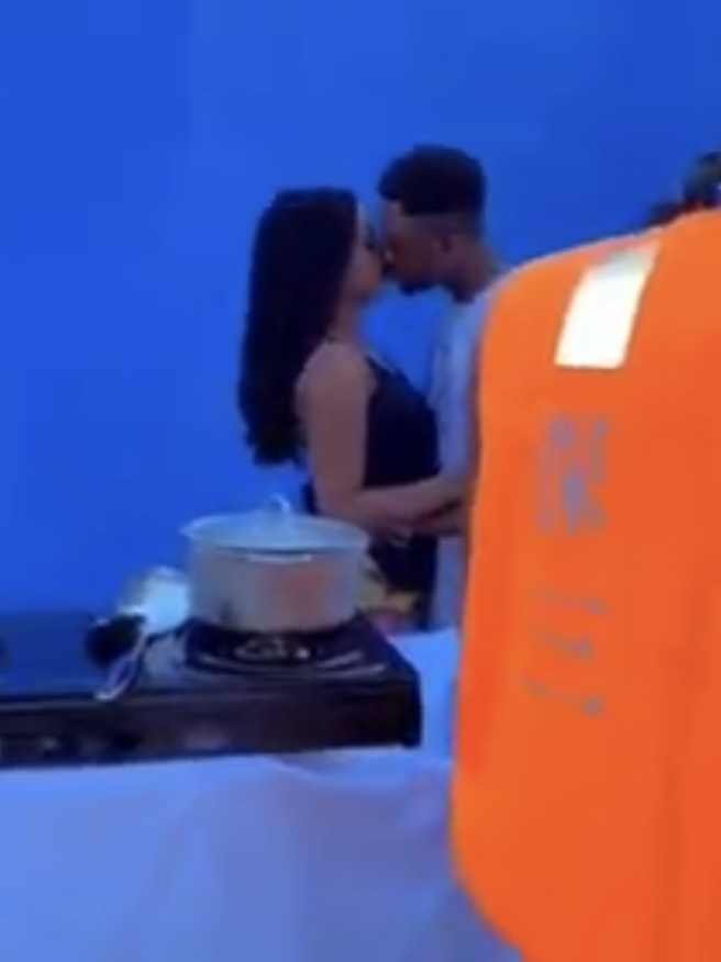 Deux personnes s’embrassant devant un mur bleu avec un plan de cuisson et une personne en gilet orange partiellement visible