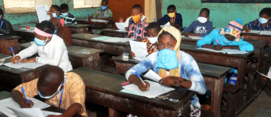 Des élèves masqués assis en rangs dans une salle de classe en train d’écrire sur des feuilles posées sur des bureaux en bois