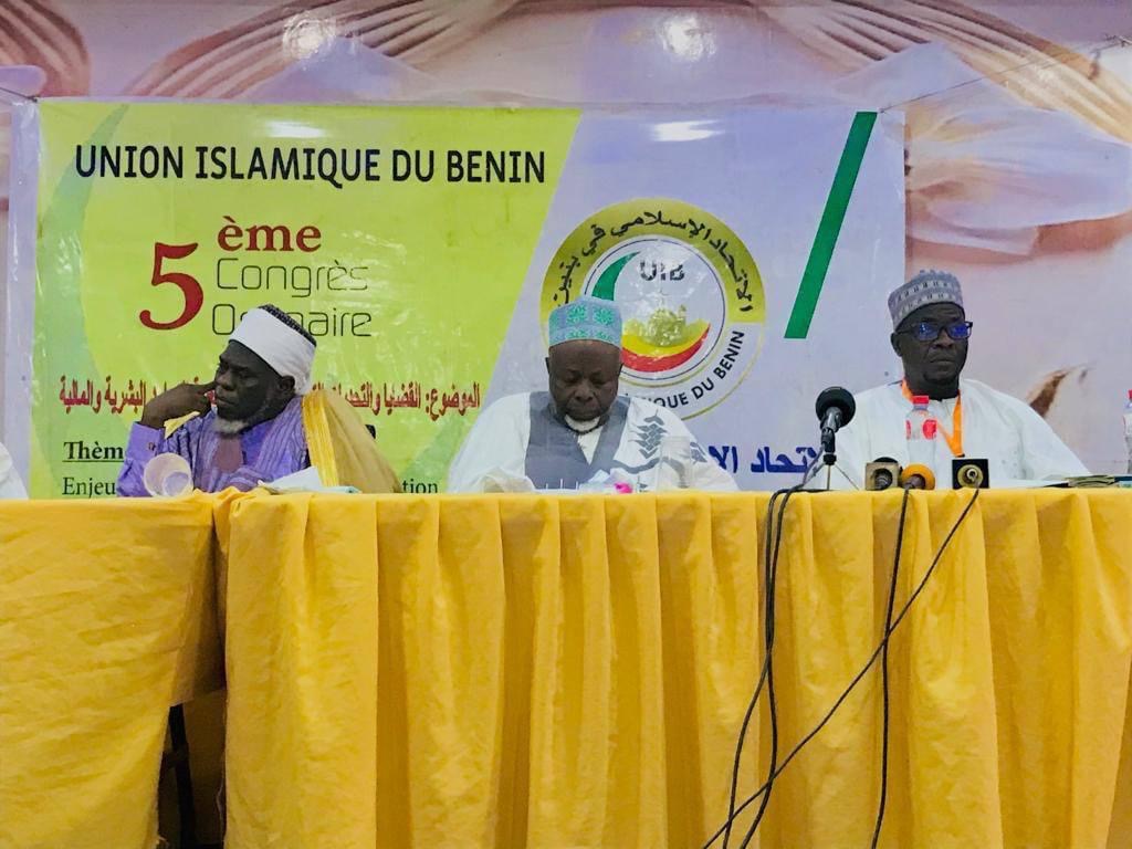 Trois hommes en tenue traditionnelle sont assis derrière une table jaune avec un panneau de congrès islamique en arrière-plan
