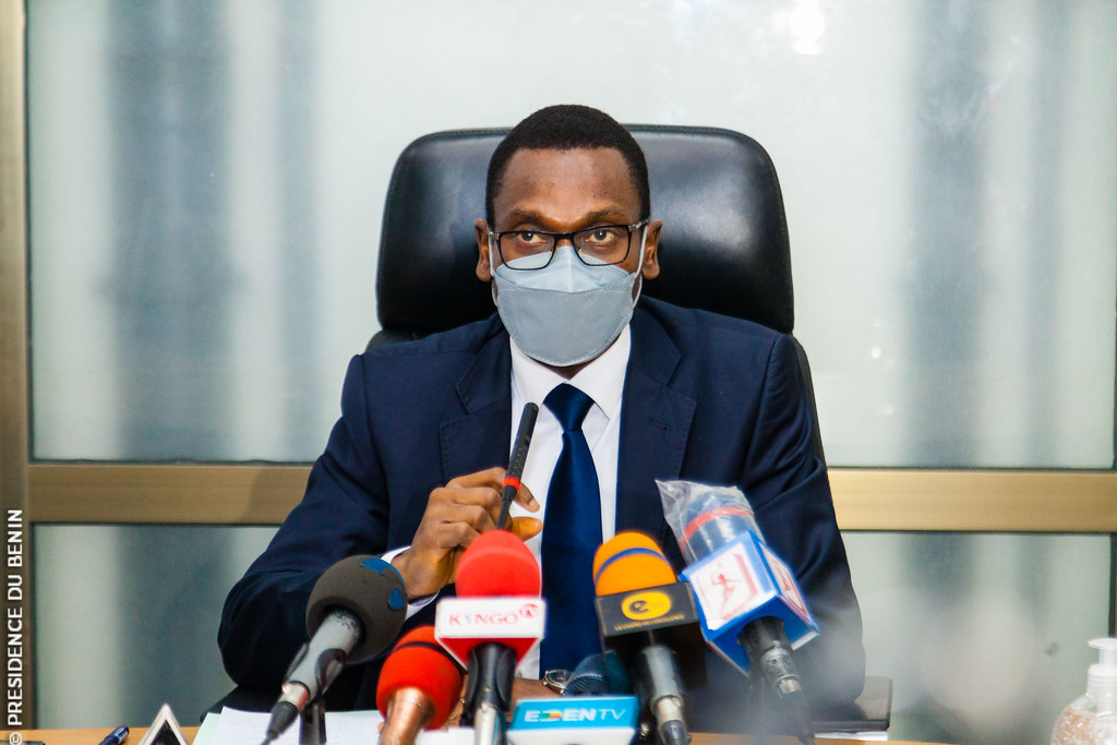 Un homme en costume et masque assis à une table avec plusieurs micros devant lui parlant dans un micro.
