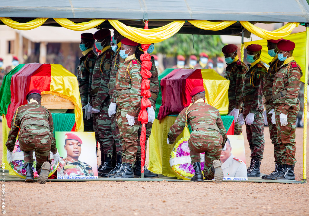 Des militaires en uniforme déposent des gerbes de fleurs devant deux cercueils sous un chapiteau décoré en jaune et rouge