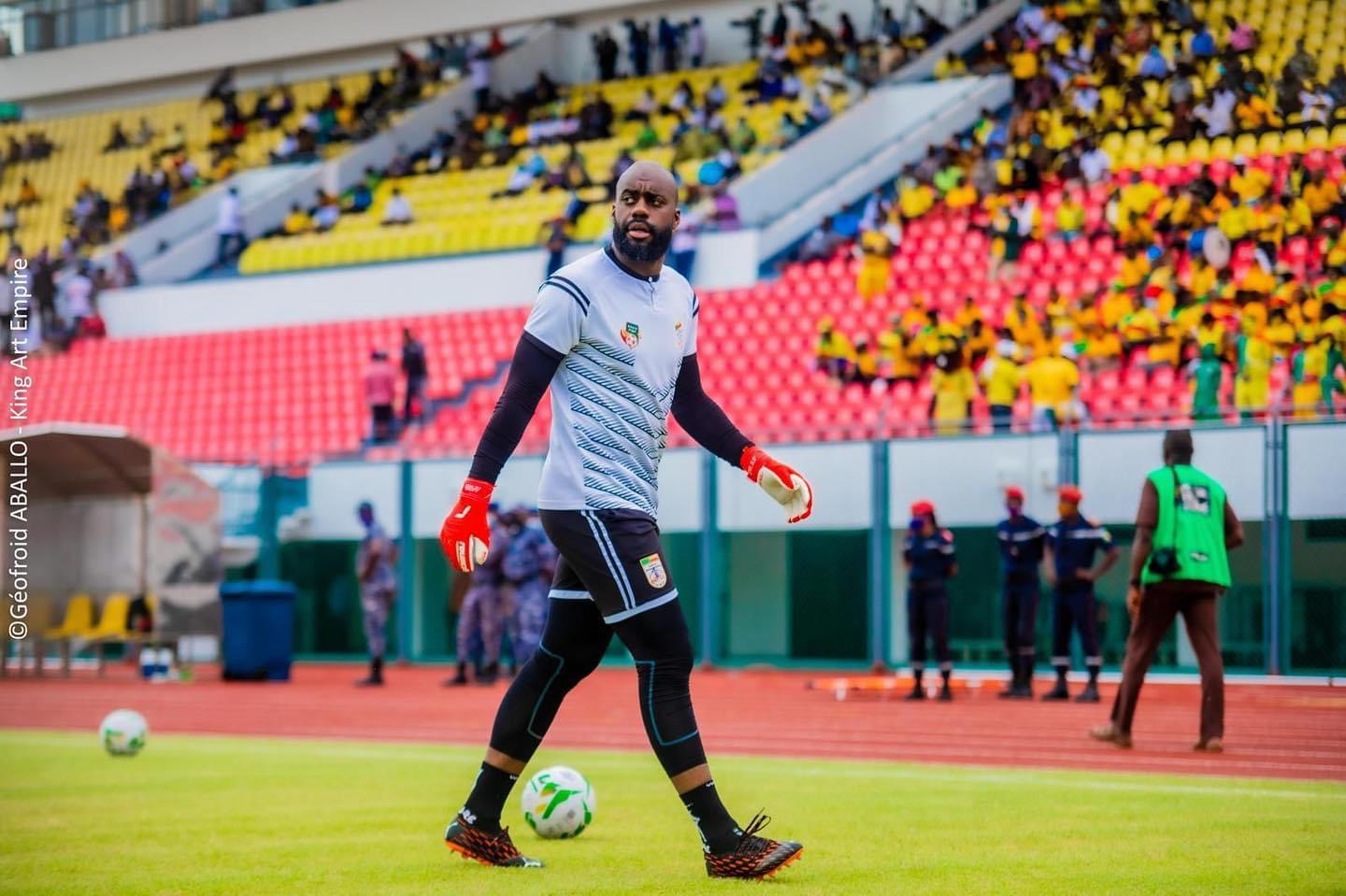 Un joueur de football en tenue d'entraînement marche sur le terrain avec un ballon au pied dans un stade rempli de spectateurs.