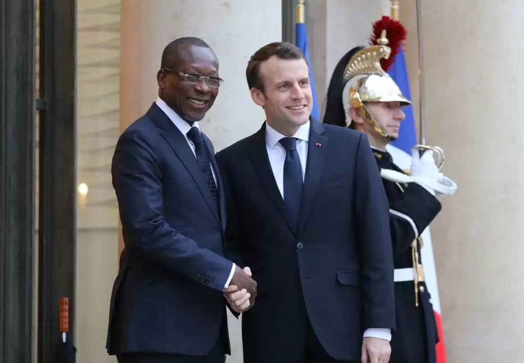 Deux hommes en costume sombre se serrent la main et sourient devant un bâtiment officiel avec un soldat en uniforme d'apparat derrière eux