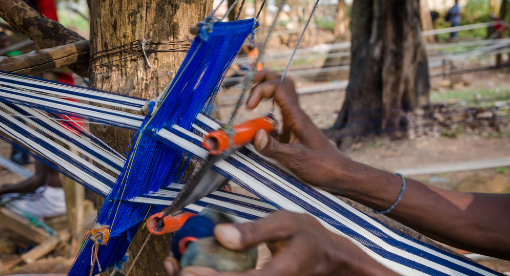Deux mains manipulent un métier à tisser artisanal avec des fils bleus et blancs dans un environnement extérieur boisé