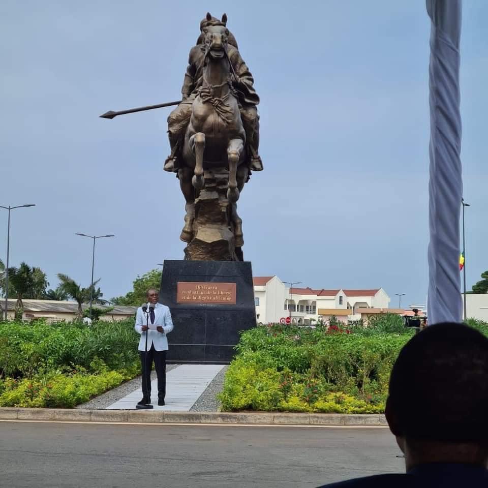 Statue équestre d un guerrier avec une lance dans un espace vert entouré de bâtiments modernes et une personne parlant au microphone devant