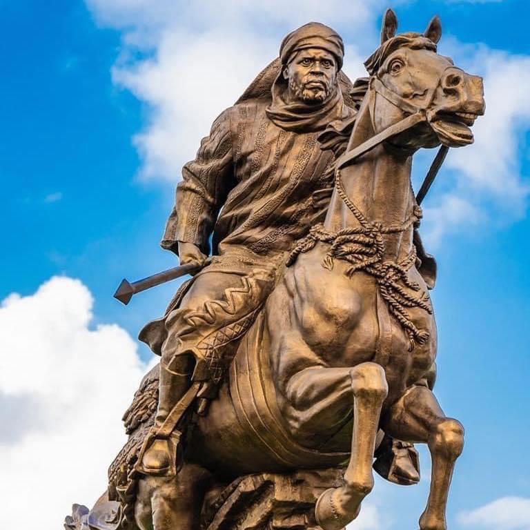 Statue en bronze représentant un homme en tenue traditionnelle à cheval brandissant un objet contre un ciel bleu partiellement nuageux
