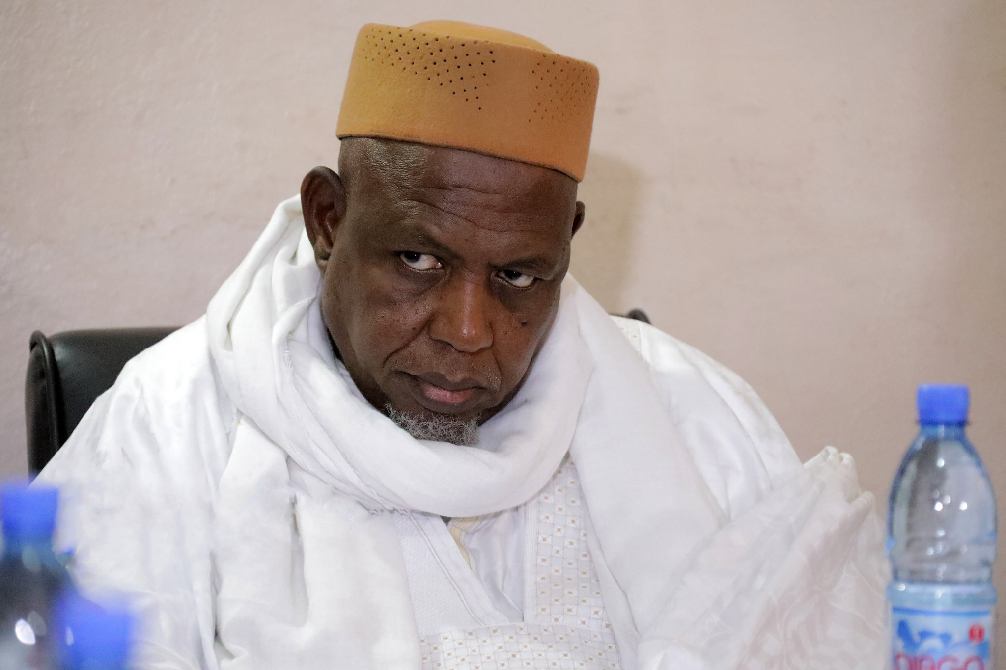 Homme portant un bonnet beige et une tenue traditionnelle blanche assis devant une table avec une bouteille d'eau bleue