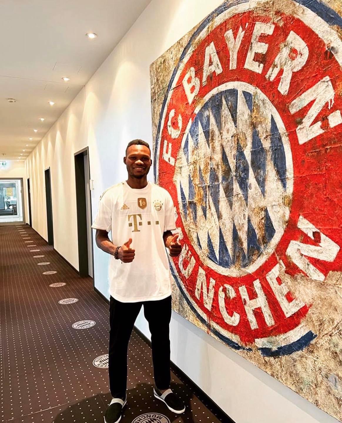 Homme debout souriant faisant un pouce levé devant un grand logo mural du FC Bayern Munich dans un couloir d'hôtel ou bureau