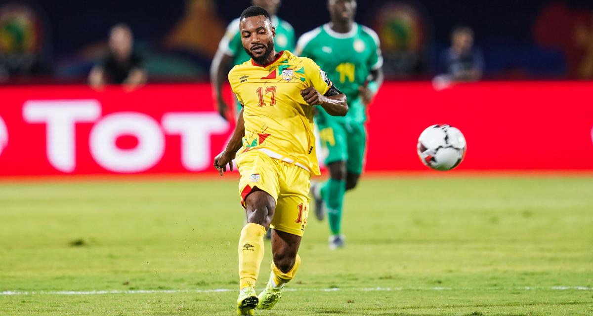 Un joueur en maillot jaune tire dans un ballon sur un terrain de football avec des adversaires en arrière-plan.