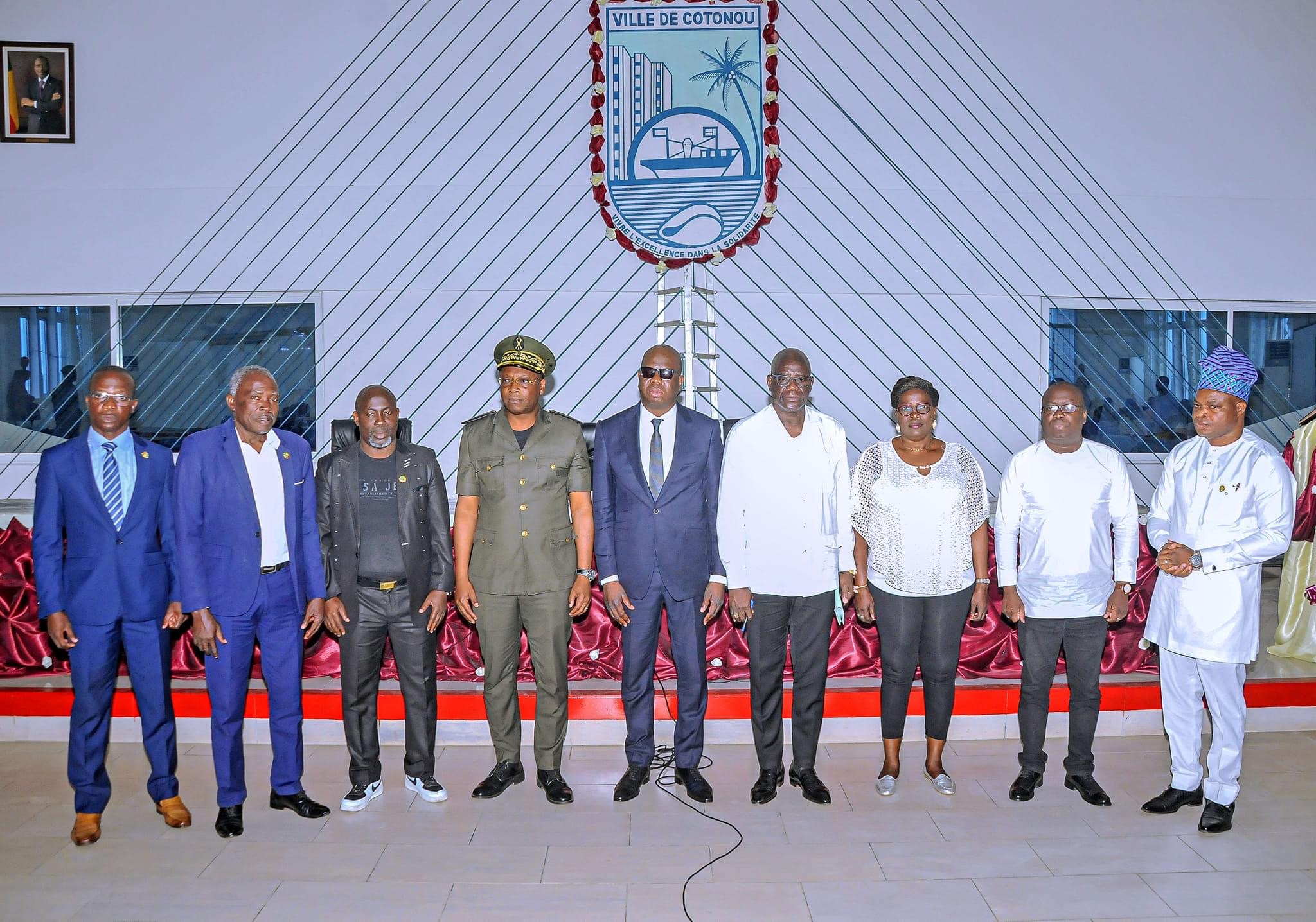 Neuf personnes debout en file devant un mur orné d’un grand blason portant l’inscription « Ville de Cotonou