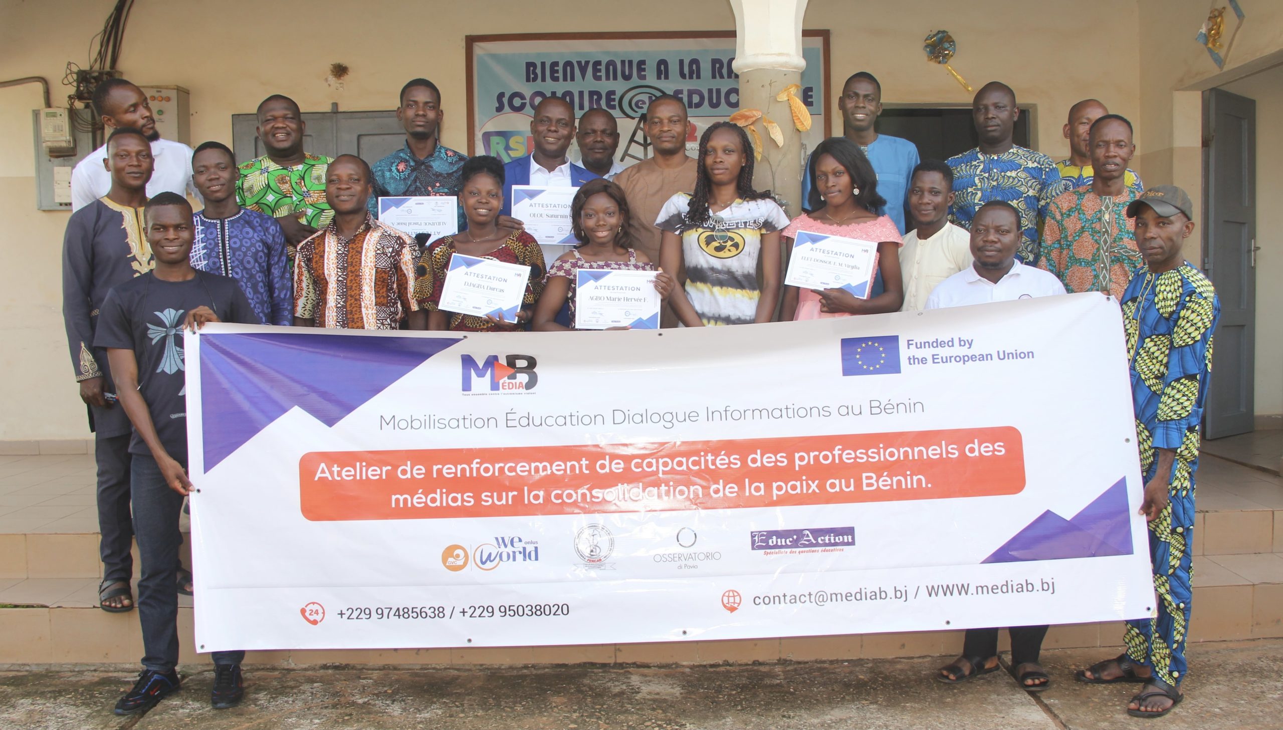 Groupe de personnes posant devant un bâtiment avec un grand panneau sur un atelier de renforcement des capacités des médias pour la paix au Bénin