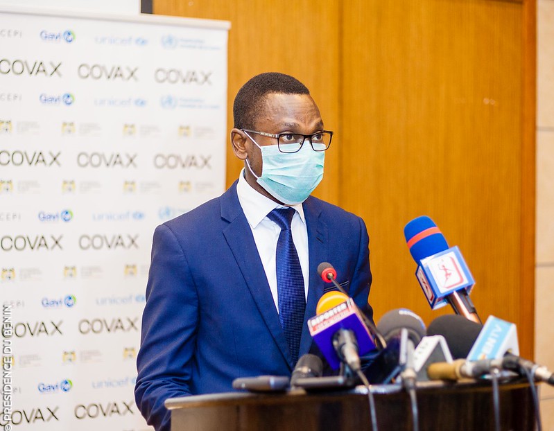 Un homme en costume bleu et masque médical parle derrière un pupitre avec plusieurs micros, logo COVAX en arrière-plan