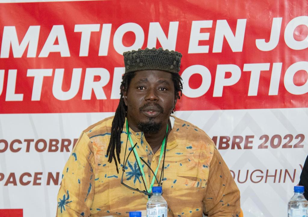 Homme à dreadlocks coiffé d’une casquette noire souriant devant une bannière rouge et blanche partiellement lisible