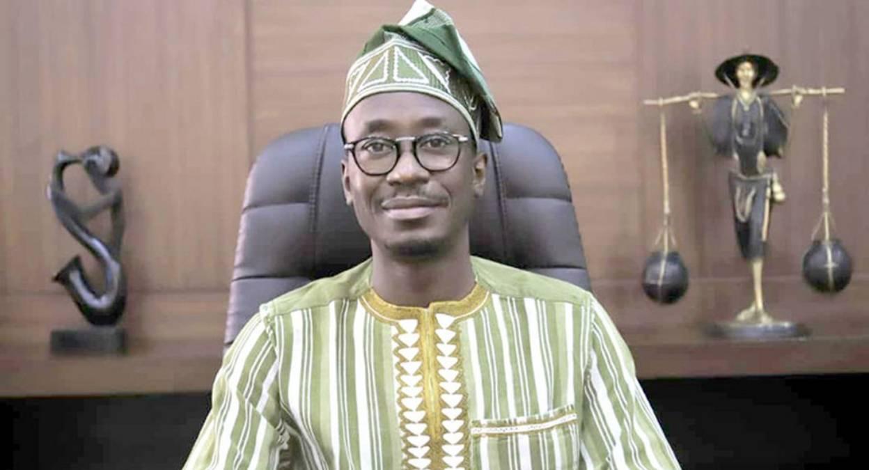 Homme portant une tenue traditionnelle rayée verte et blanche avec un chapeau assorti assis devant un bureau avec des sculptures en arrière-plan
