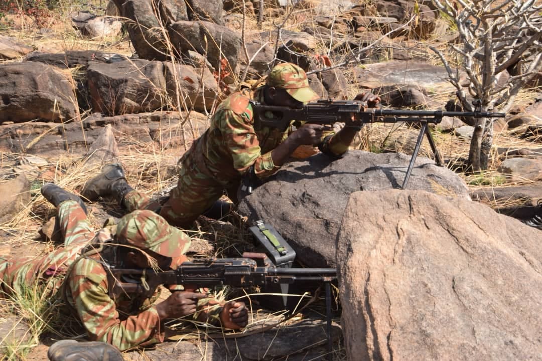 Deux soldats en tenue camouflage armés sont positionnés au sol derrière un rocher dans un environnement sec et rocailleux