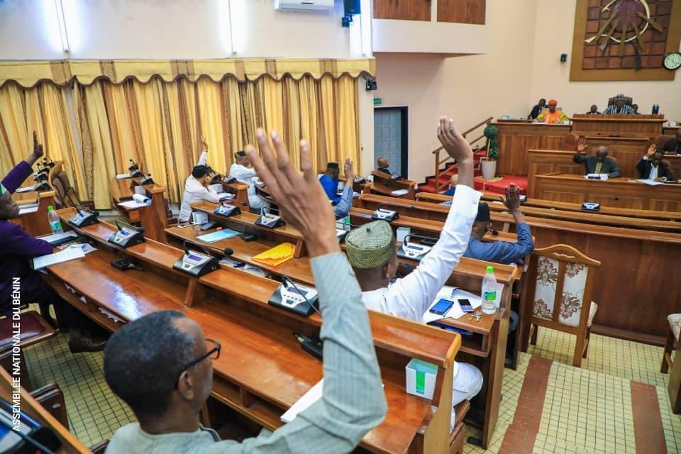 Plusieurs personnes lèvent la main dans une salle de réunion officielle équipée de bancs en bois et d'un pupitre central.