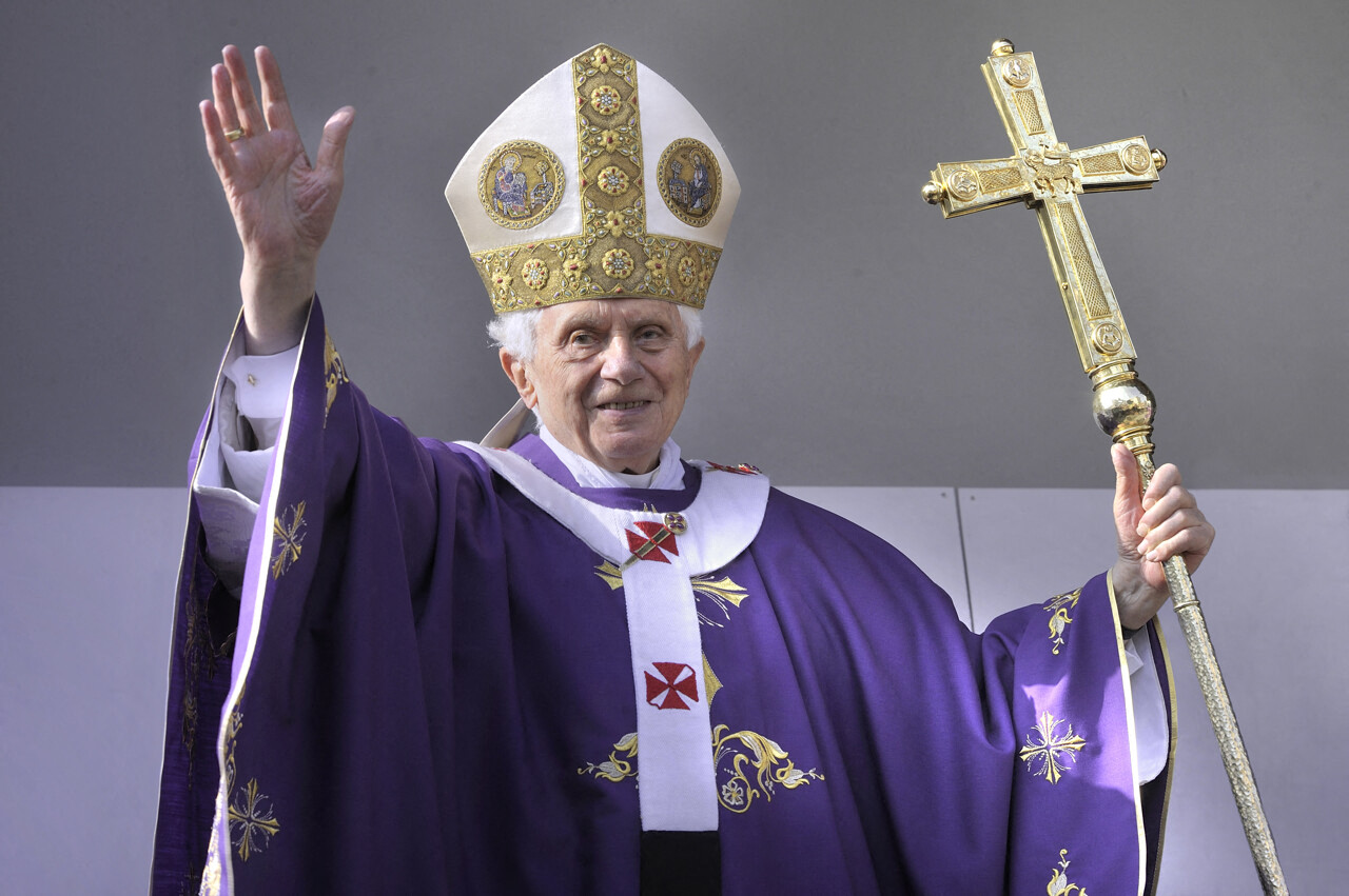 Un homme âgé en tenue ecclésiastique pourpre avec une mitre dorée lève la main droite et tient une grande croix dorée de la main gauche