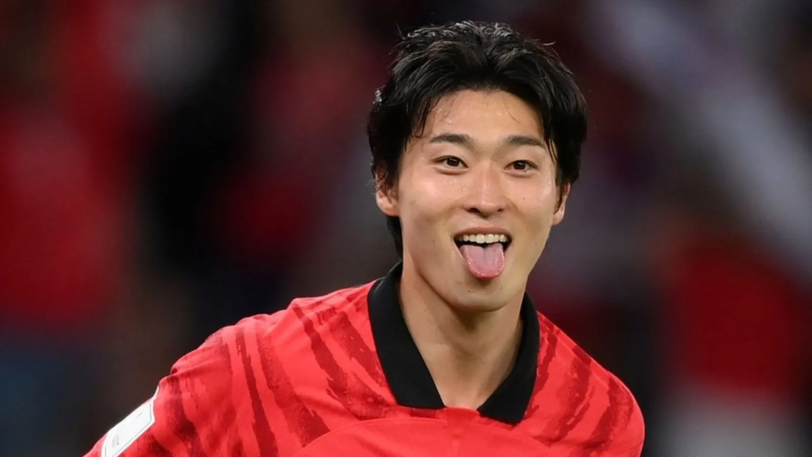 Joueur de football asiatique en maillot rouge tire la langue en souriant sur un fond flou rouge et noir