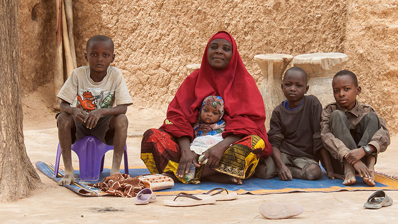 Une femme voilée en rouge assise sur un tapis avec quatre enfants dans un environnement extérieur sec et simple