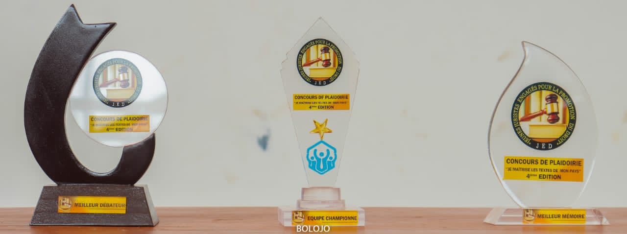 Trois trophées alignés sur une surface boisée avec inscriptions dorées et logos visibles sur un mur clair en arrière-plan