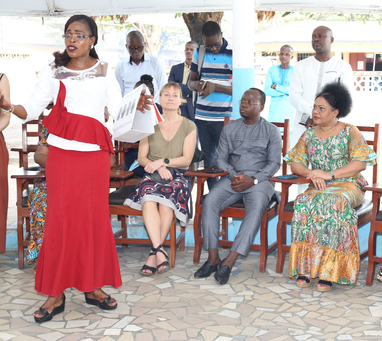 Une femme en robe rouge parle devant un petit groupe assis attentivement sous un abri ouvert